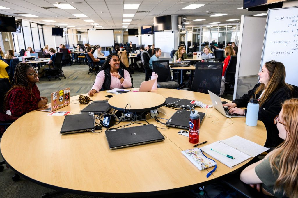 Large classroom with multiple round tables with laptops and notebooks where students are seated, having a discussion.