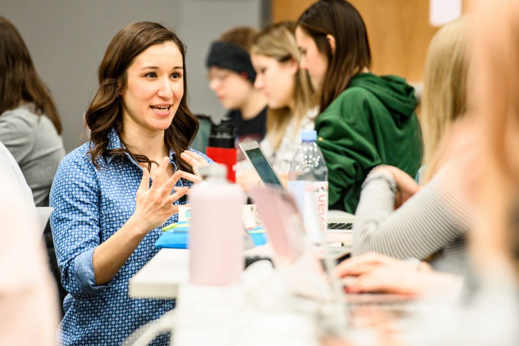 A woman is seated at a table in a classroom setting, engaged in discussion, with laptops, notebooks, and other students around her.