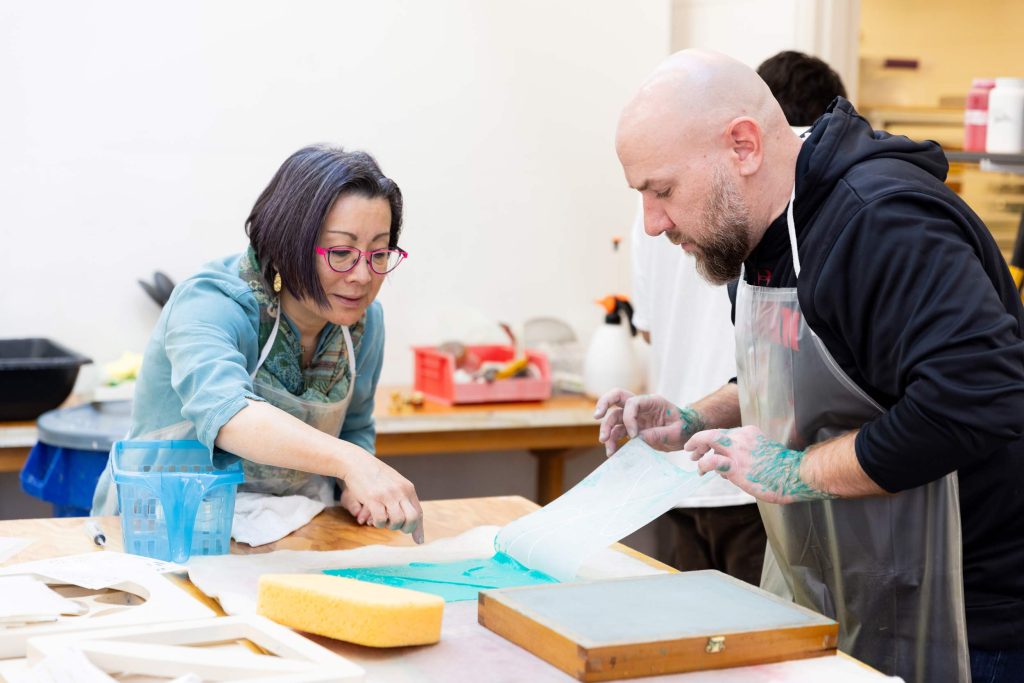 Two people wearing aprons work together at a table making paper in a studio setting.