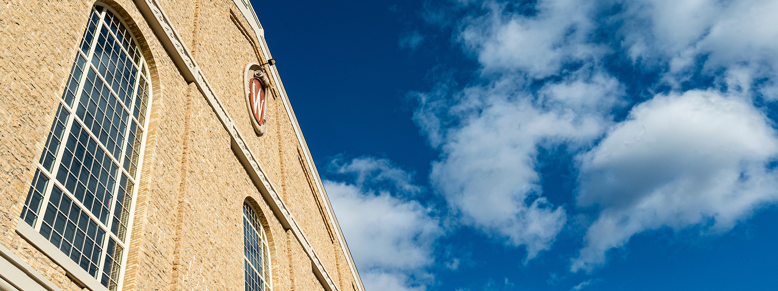 Upward view of the exterior of the UW Field House with tall arched windows and the red 'W crest against a bright blue sky with scattered white clouds.
