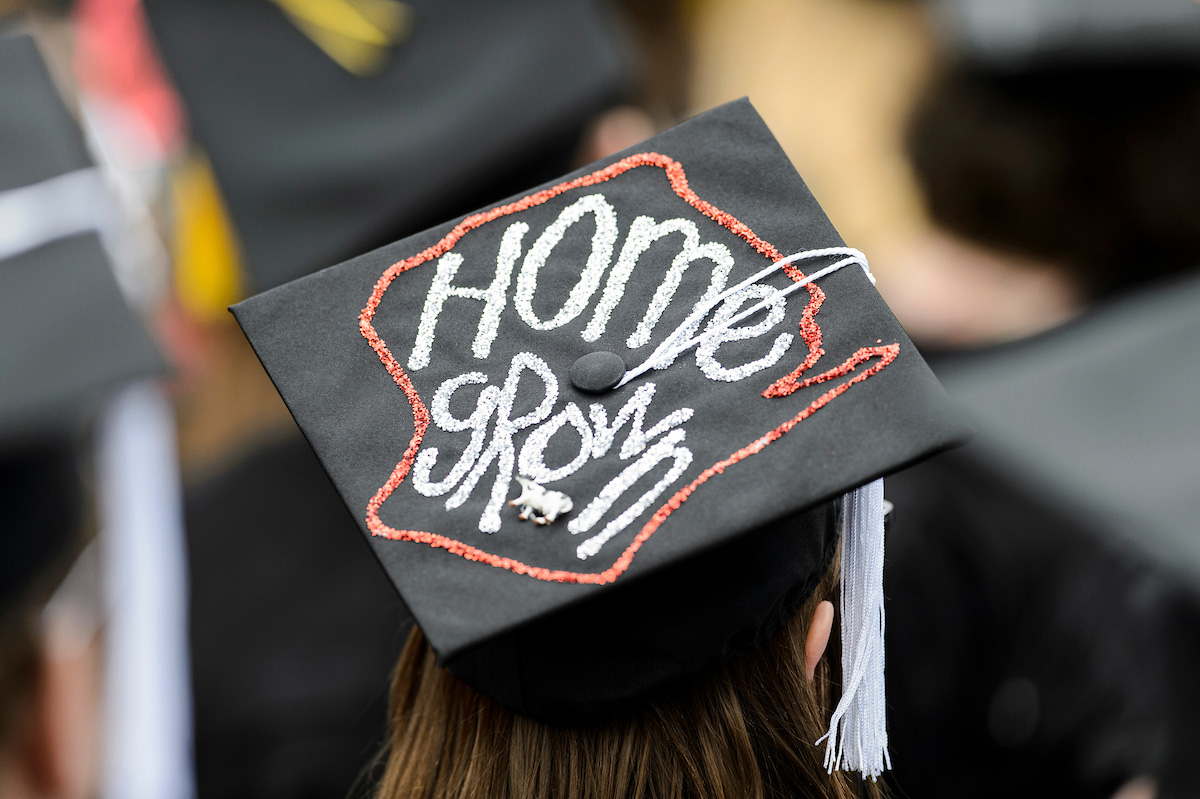 A mortarboard decorated with an outline of the state of Wisconsin in red glitter. Inside is hand printed 'Home grown' in silver glitter.