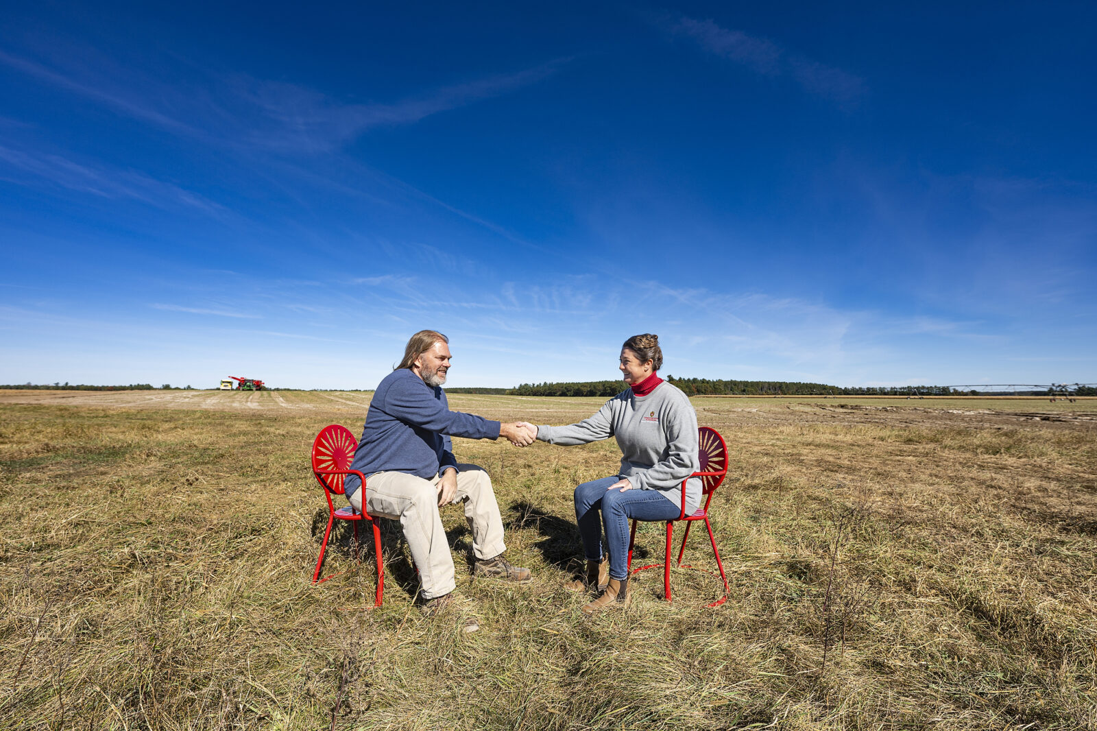 Two people sit in Badger red metal chairs in the middle of a brown potato field under a blue sky. They reach out and shake hands with each other.