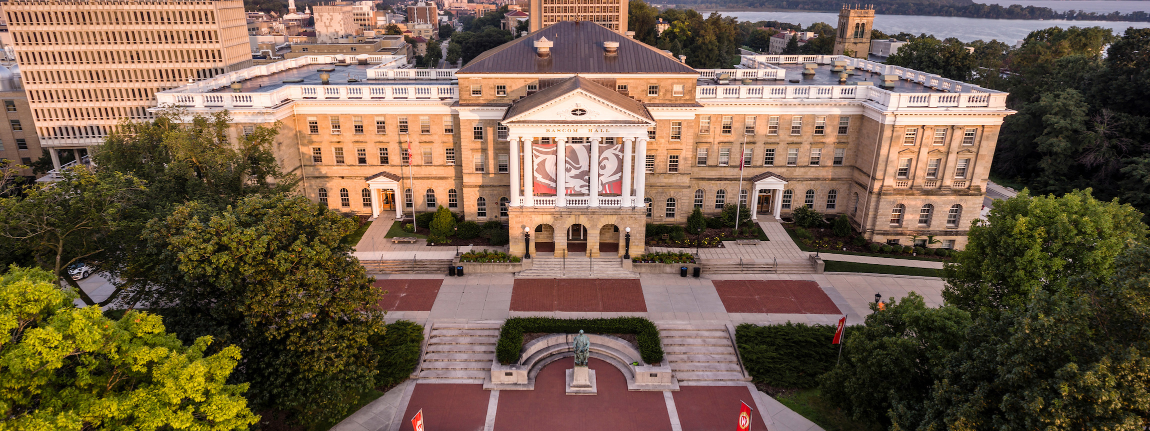 A wide drone shot of Bascom Hall at dawn, illuminated by warm morning sunlight with Lake Mendota visible in the distance and a large red-and-white Bucky Badger banner hanging between the central white columns.