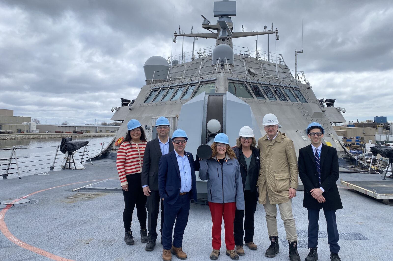 Five people in hard hats stand in a row and smile to the camera while standing on the deck of a U.S. Navy combat ship on an overcast day.