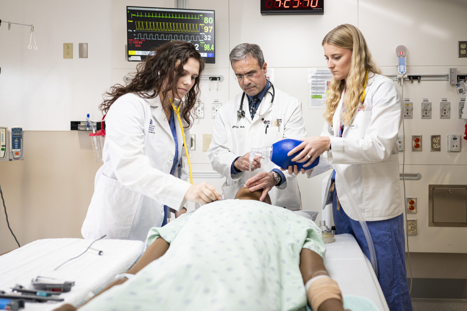 Doctor demonstrates procedure on medical dummy lying on a gurney to two medical students, all three wearing white lab coats